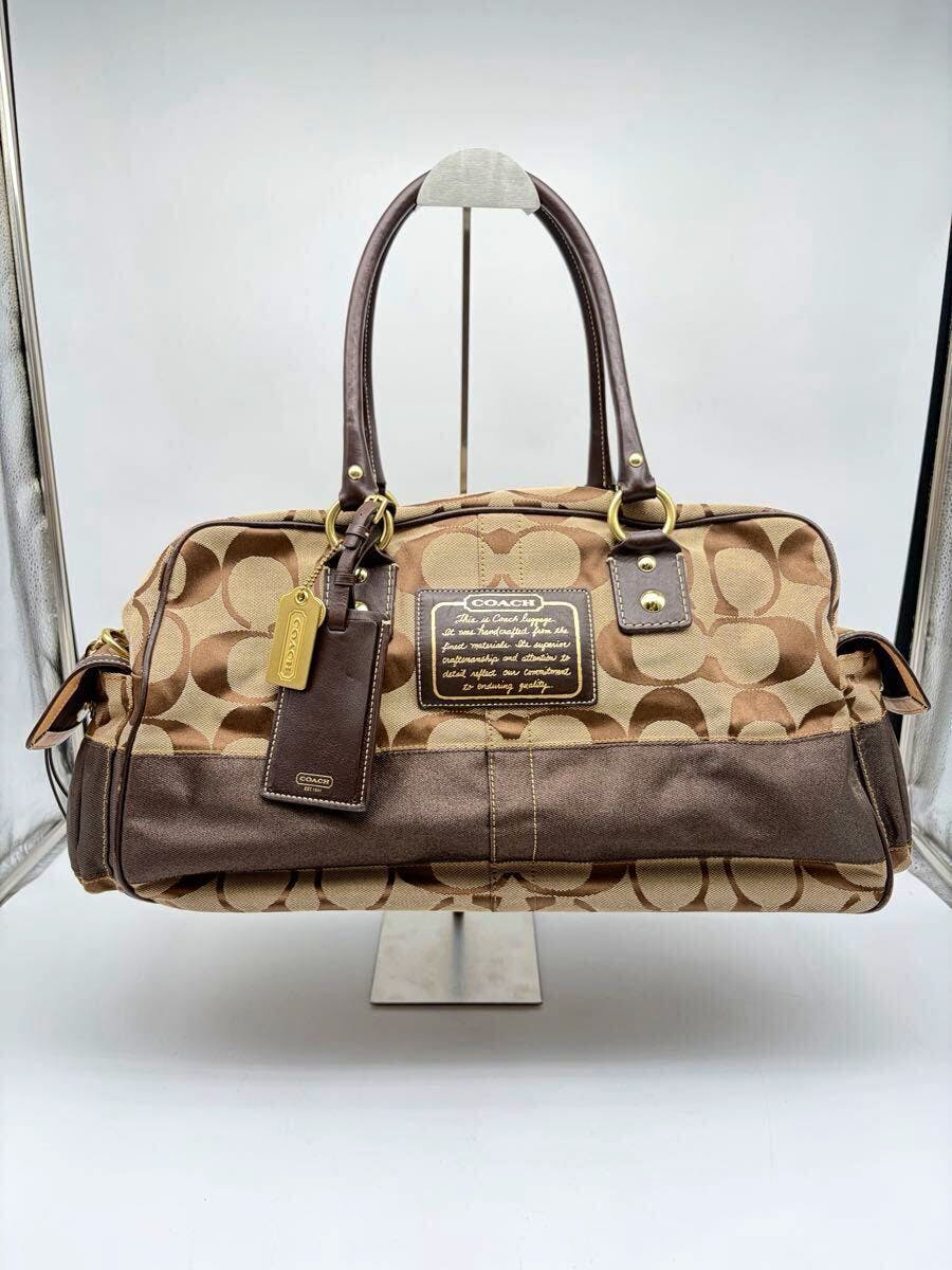 Front view of a brown Coach signature canvas Boston handbag with dark leather handles and a Coach logo patch.