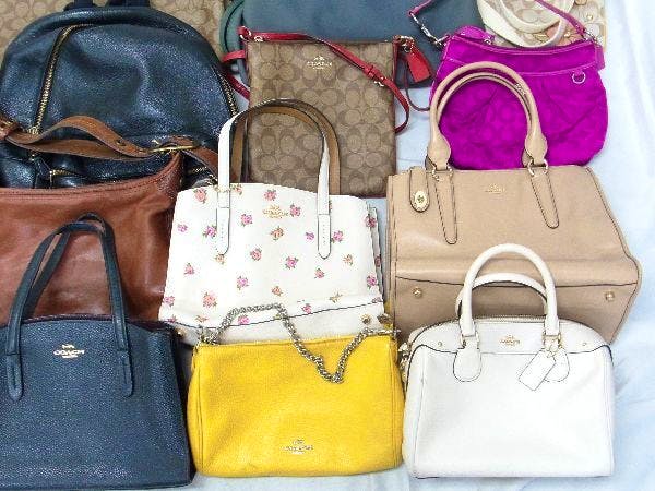 A close photo of multiple COACH handbags and tote bags in various colors arranged on a light surface.