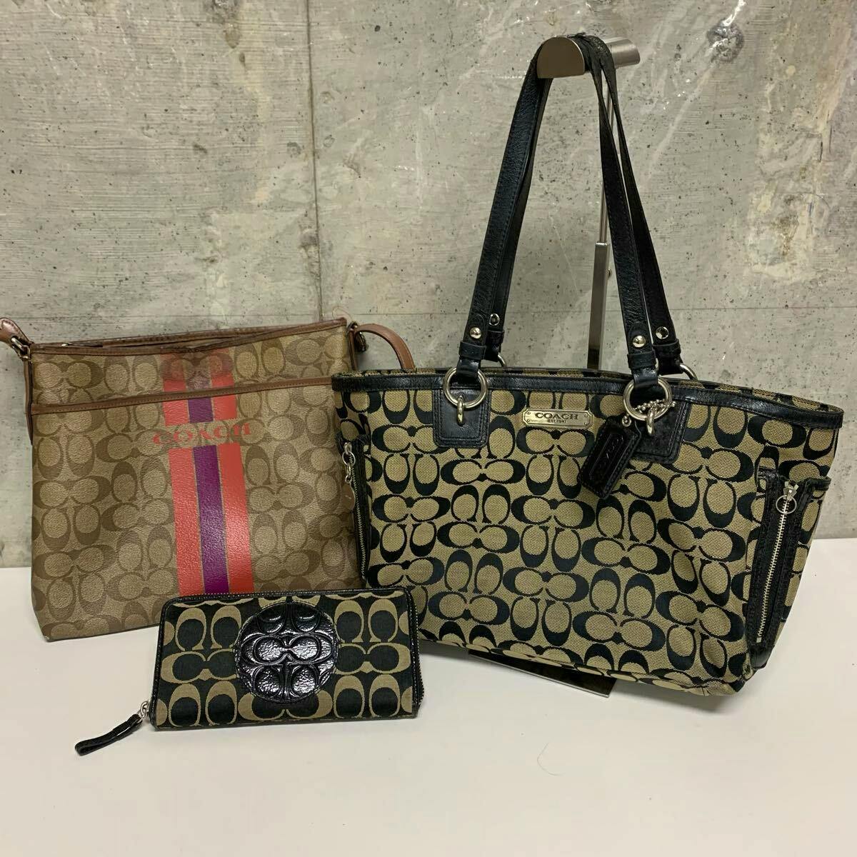 Three Coach items: two signature canvas bags (a tote and a shoulder bag) and a zip-around long wallet displayed on a white surface against a concrete wall.