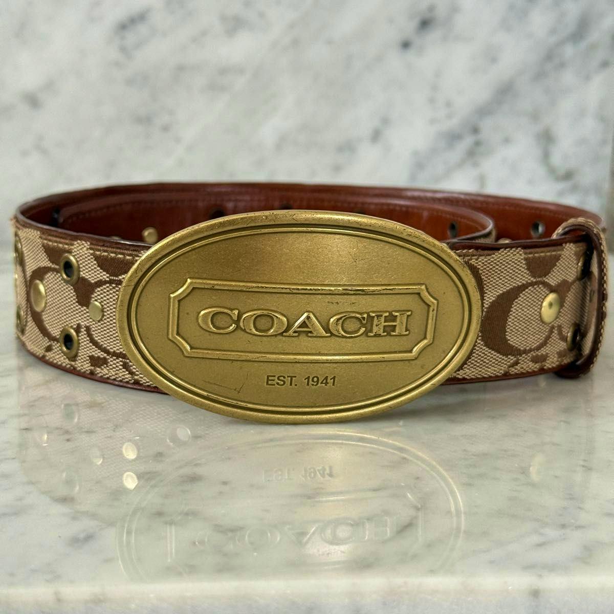 Front view of a COACH brown signature canvas and leather belt with an oval metal buckle stamped "COACH EST. 1941" and decorative metal studs.