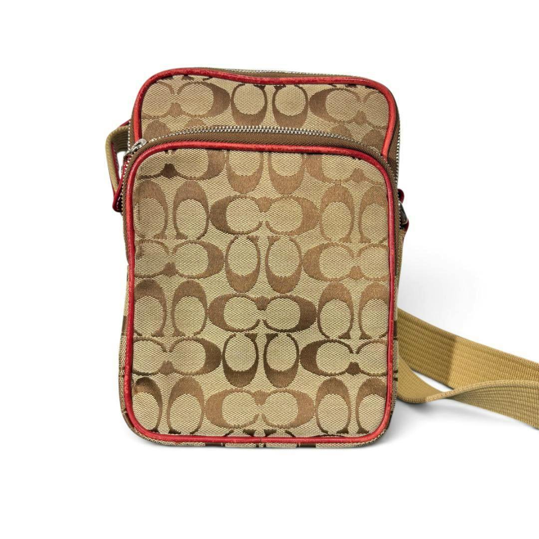 Front view of a beige Coach signature canvas crossbody shoulder bag with red trim and a tan adjustable strap.