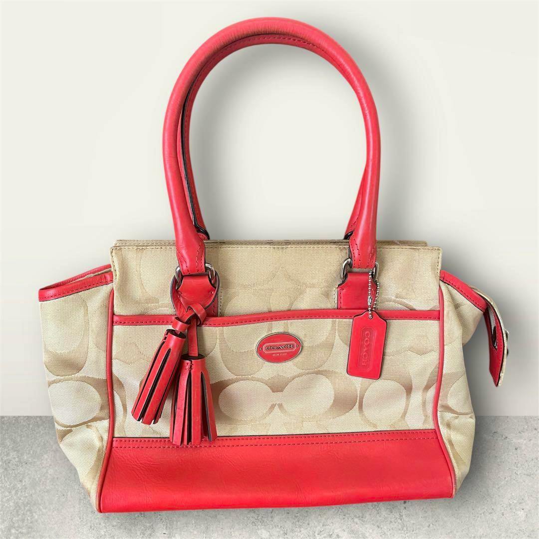 Beige Coach signature canvas tote bag with orange leather trim, handles, tassel charms, and a Coach logo tag on a neutral background.