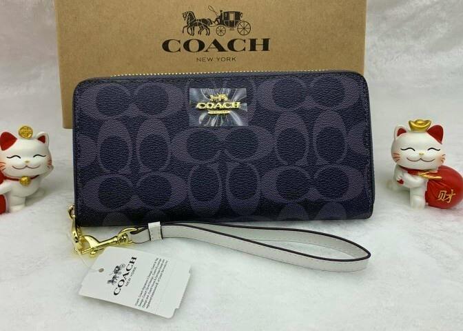 Front view of a COACH signature canvas long zip-around wallet with a gold-tone logo plaque and detachable wrist strap, shown with a COACH box behind it.