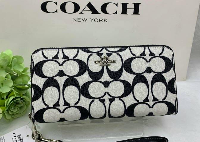 Front view of a COACH long wallet in black-and-white signature pattern with a metal Coach logo, displayed in front of a COACH box.