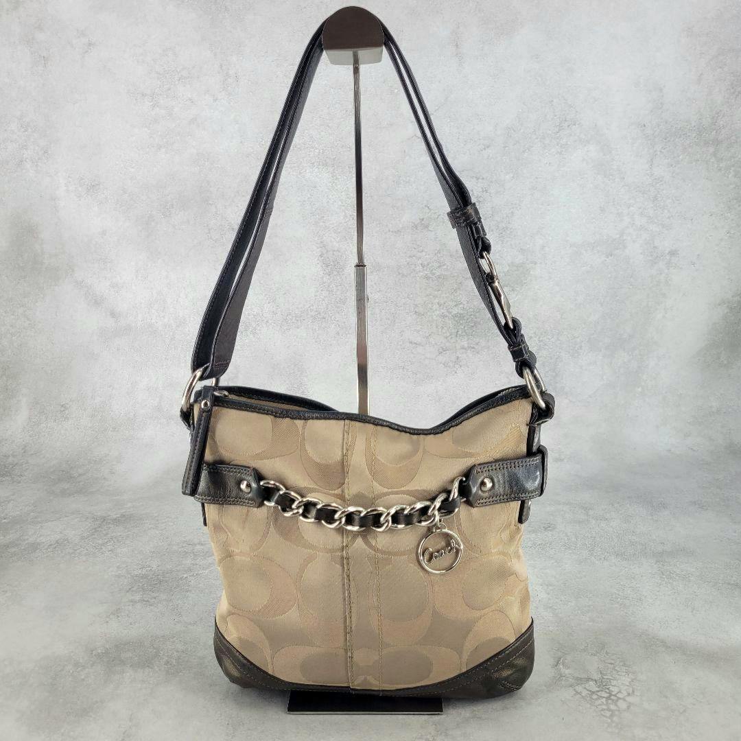 Front view of a beige Coach signature canvas one-shoulder bag with dark leather trim, a silver chain accent, and a round Coach charm.