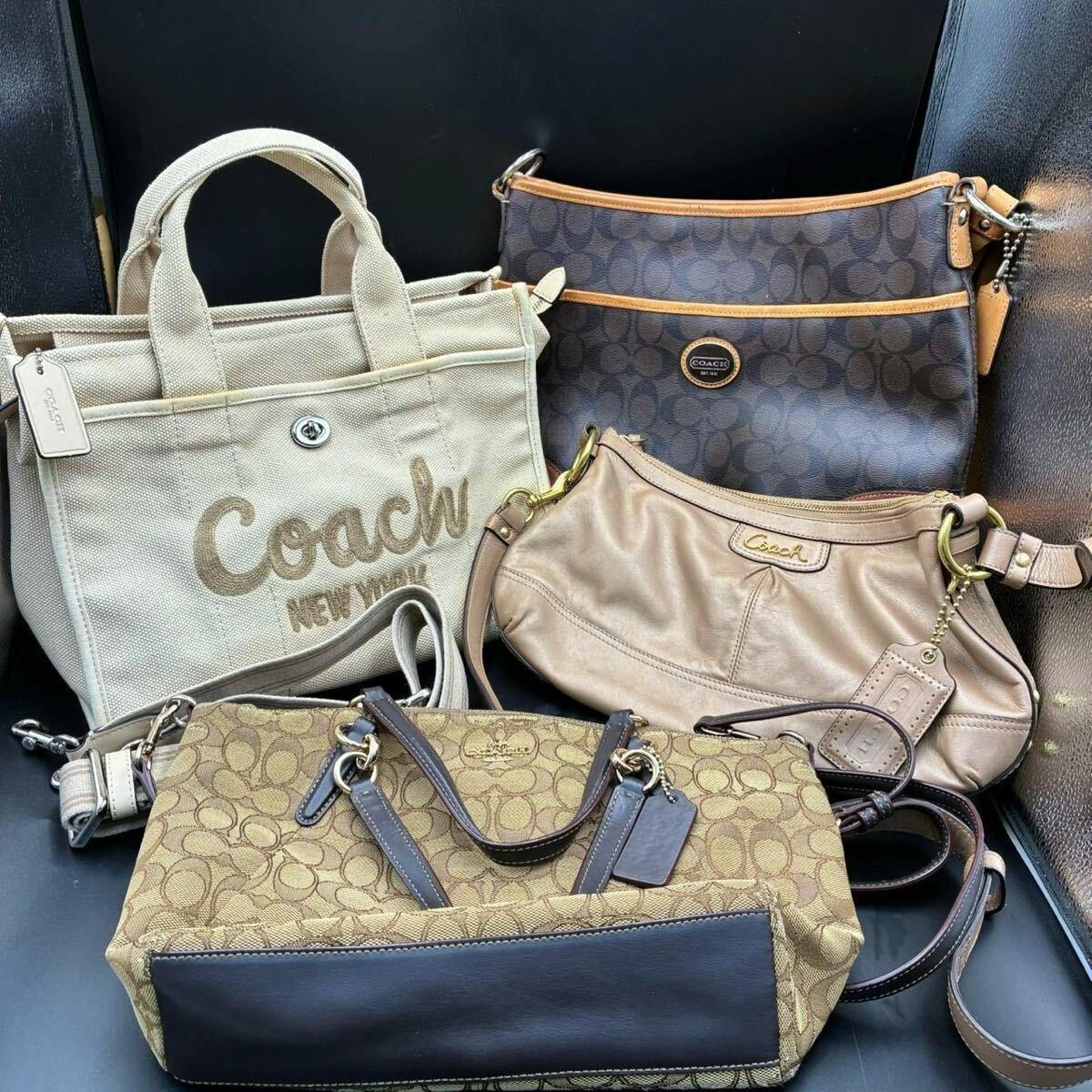 Four COACH bags arranged together including a beige canvas 'Coach New York' tote, two signature monogram bags, and a small tan leather shoulder bag.