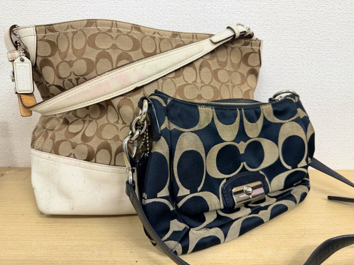 Two COACH signature canvas bags, a beige tote and a navy logo shoulder bag, shown resting on a wooden surface.