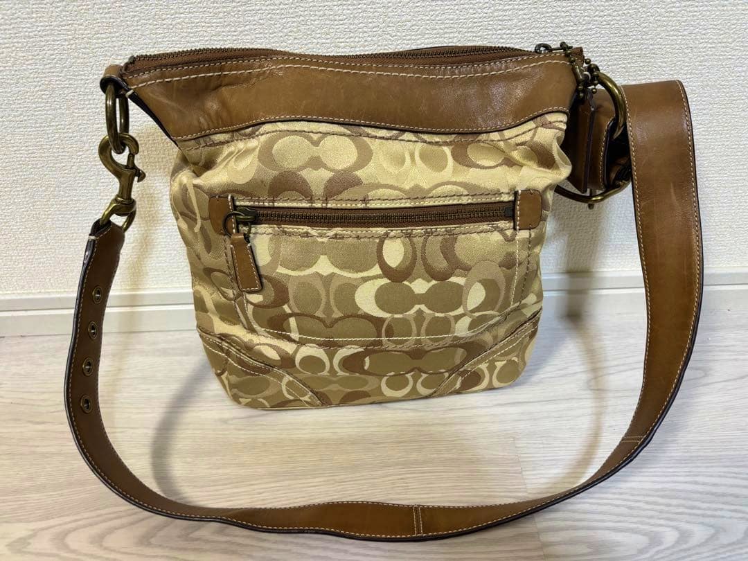 Beige COACH signature canvas shoulder bag with brown leather trim, front zip pocket and adjustable leather strap shown front-facing on a floor background.
