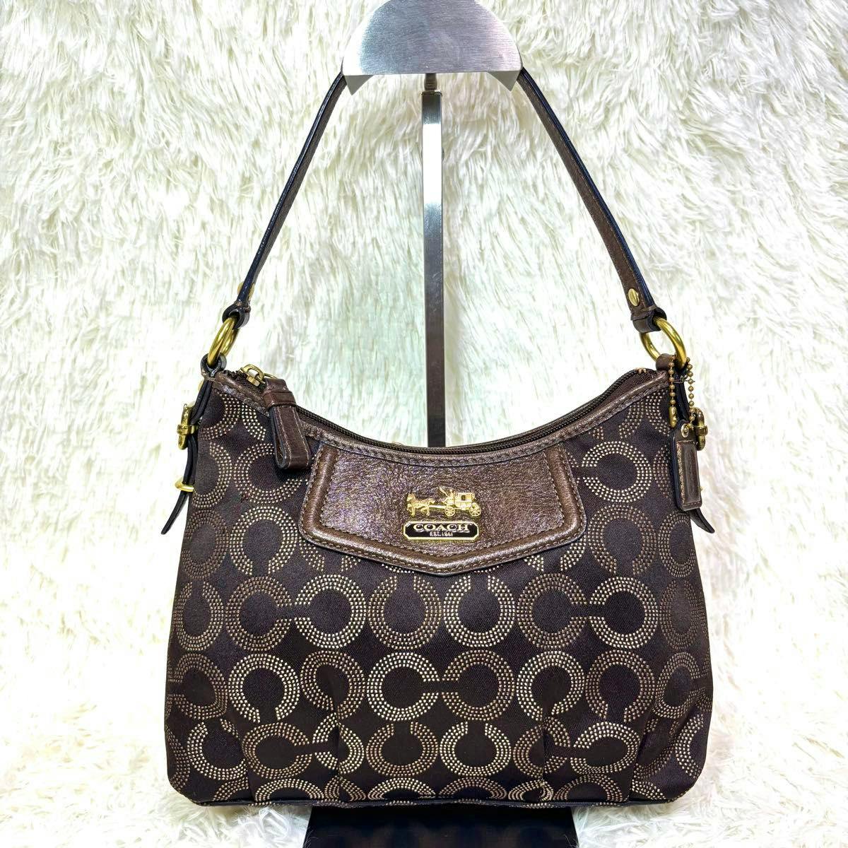 Front view of a brown COACH signature canvas shoulder bag with circular 'C' pattern, leather trim, and a short shoulder strap displayed on a stand.