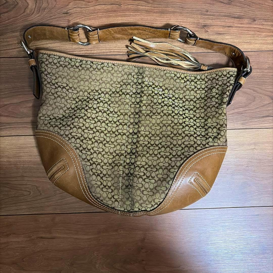 COACH signature canvas shoulder bag with leather trim, strap, and tassel lying flat on a wooden floor.
