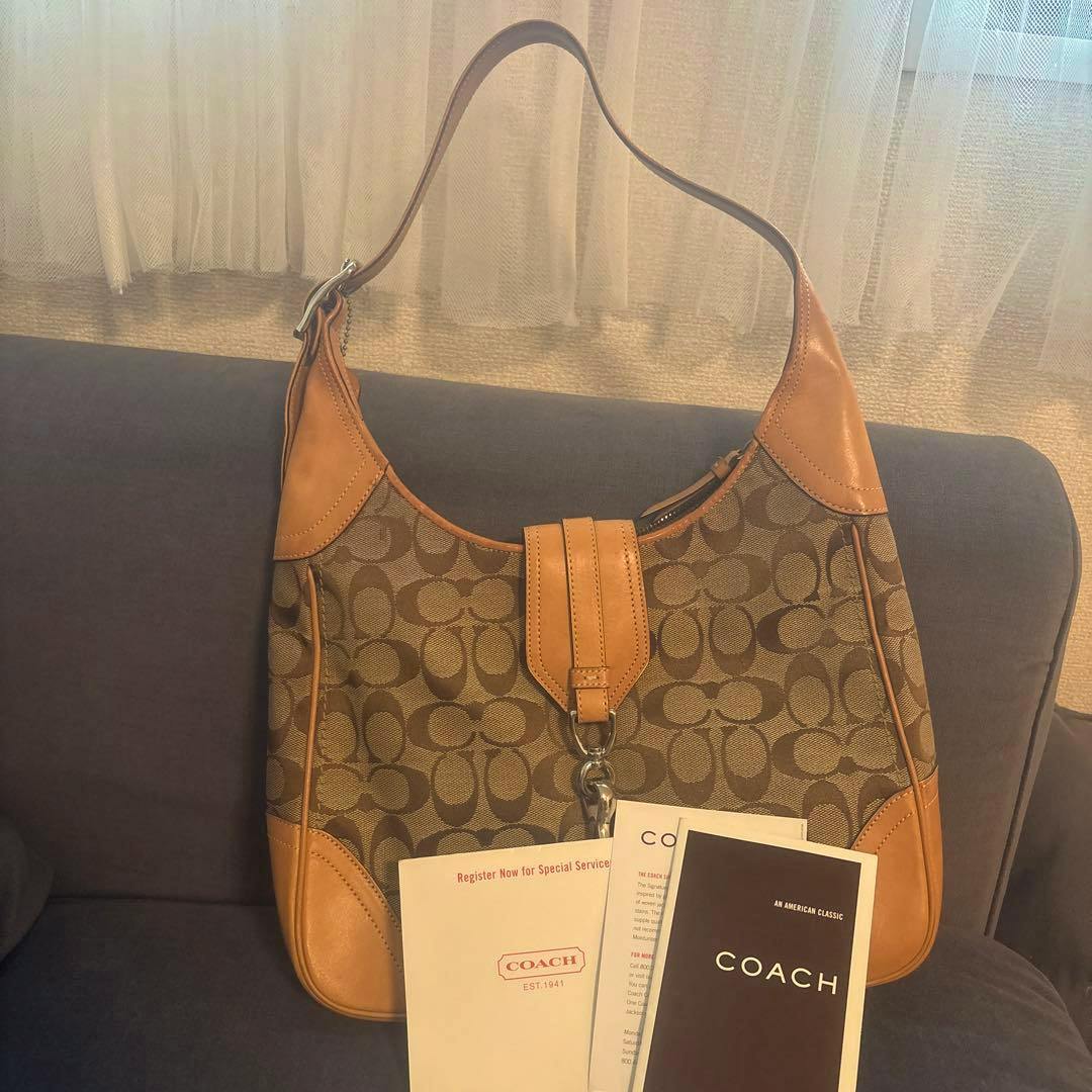 Front view of a Coach signature canvas shoulder bag with tan leather trim and Coach paperwork placed in front.