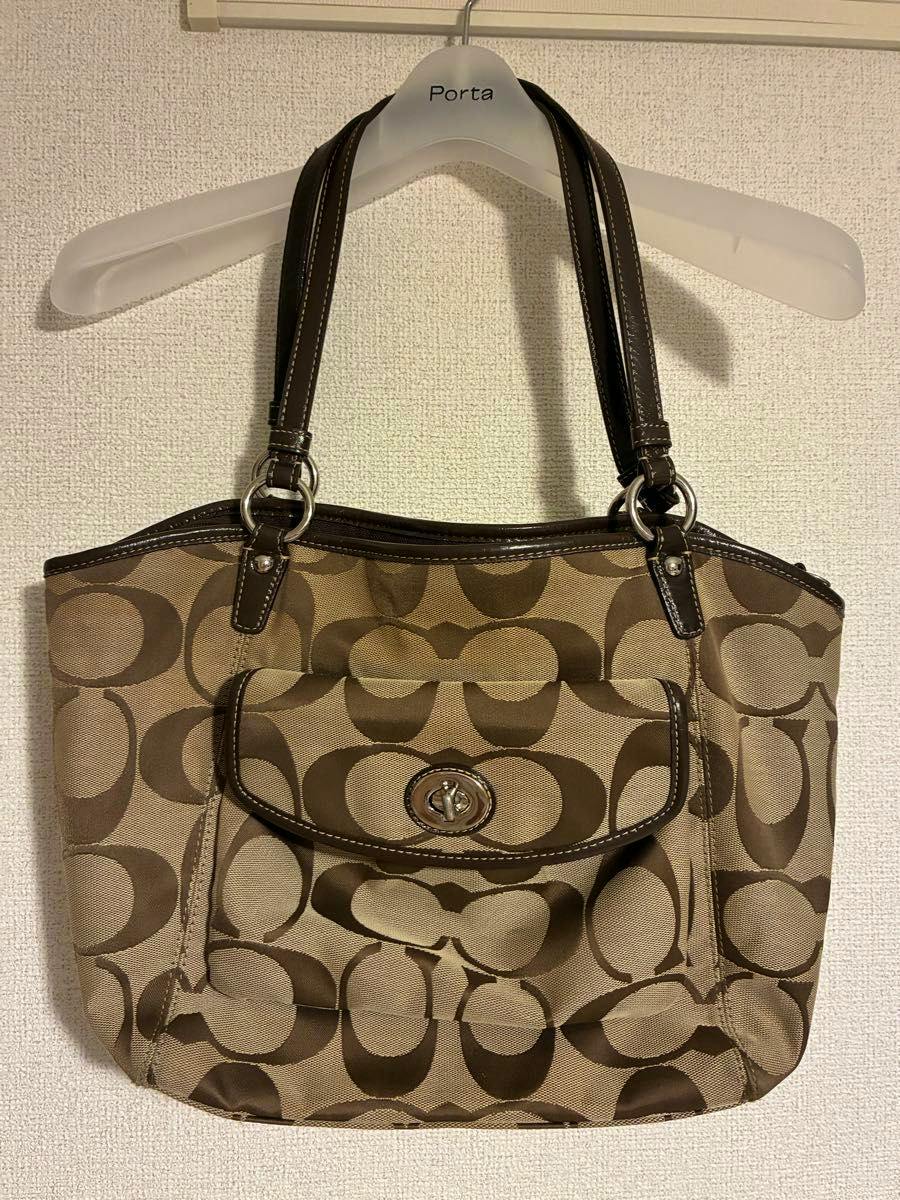 Front view of a COACH signature canvas tote bag hanging on a hanger against a textured wall.