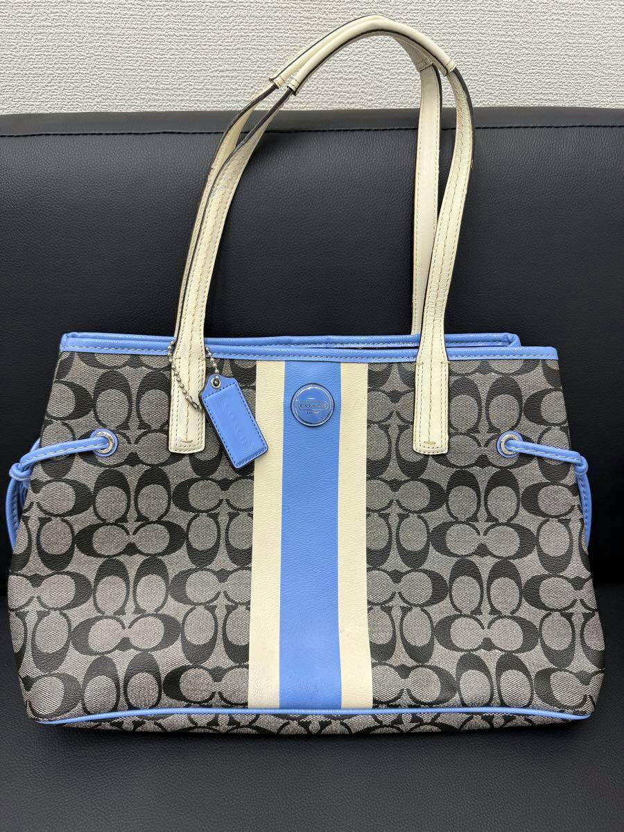 Front view of a COACH signature canvas tote bag showing blue and cream vertical center stripes and cream-colored handles on a black background.