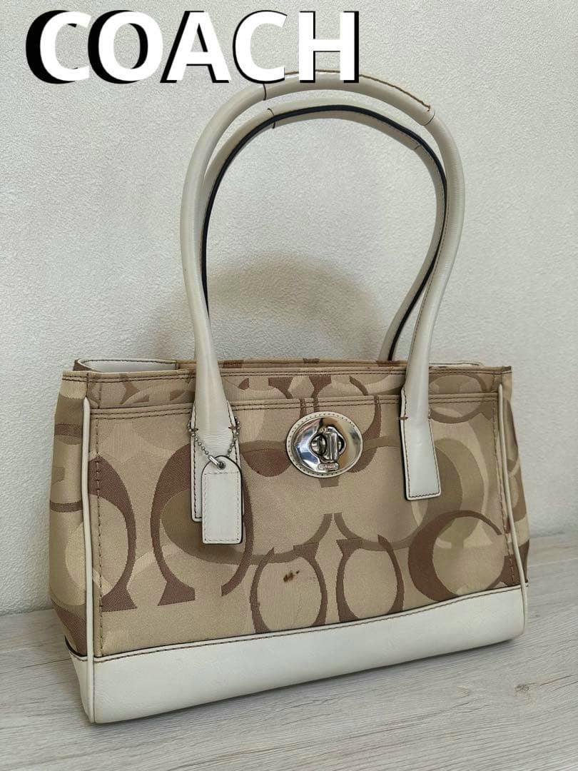 Beige Coach signature canvas tote bag with white leather trim and double handles, shown front-facing on a light surface.