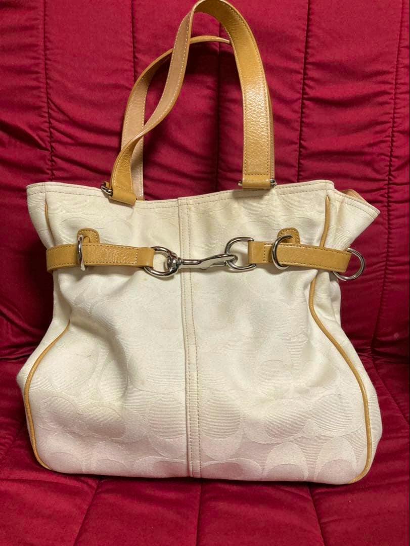 Front view of a white COACH signature canvas tote bag with tan leather handles and silver metal bit hardware against a red background.