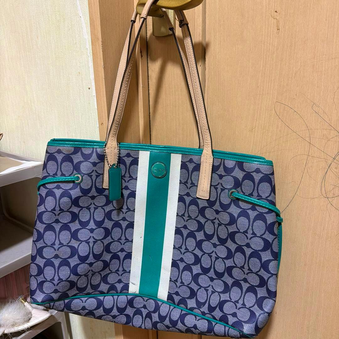 Front view of a COACH signature canvas tote bag with teal center stripe and leather handles hanging on a hook.