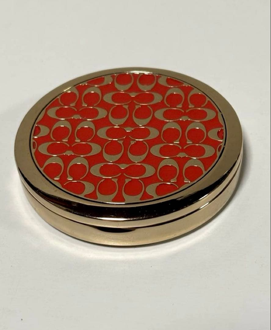 Round COACH compact mirror showing the red signature pattern on the top of the compact mirror.