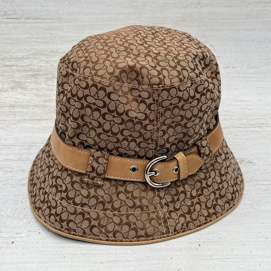 Brown Coach signature-pattern cotton bucket hat with a tan belt and silver buckle shown on a light background.