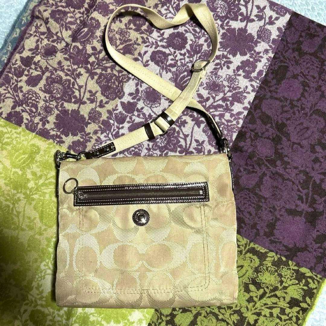 Beige Coach signature canvas crossbody bag with a front zip pocket and adjustable strap laid on patterned fabric.