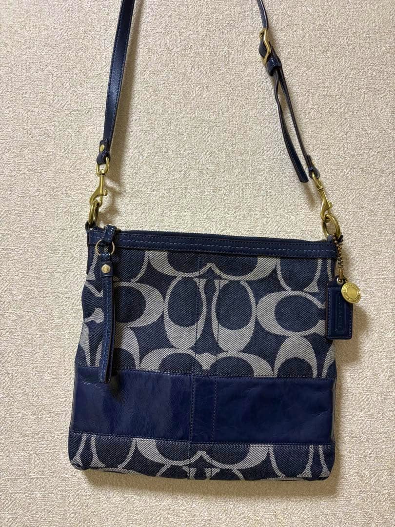 Front view of a Coach signature canvas denim shoulder bag with leather trim and adjustable strap hanging on a wall.