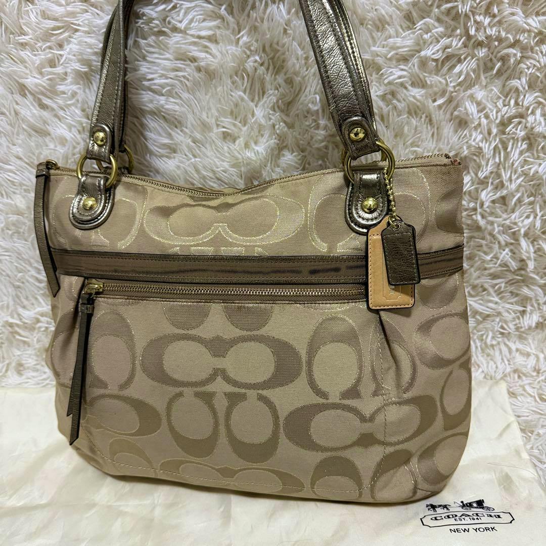 Front view of a gold COACH tote bag with signature 'C' pattern, metallic straps, and a front zip pocket.