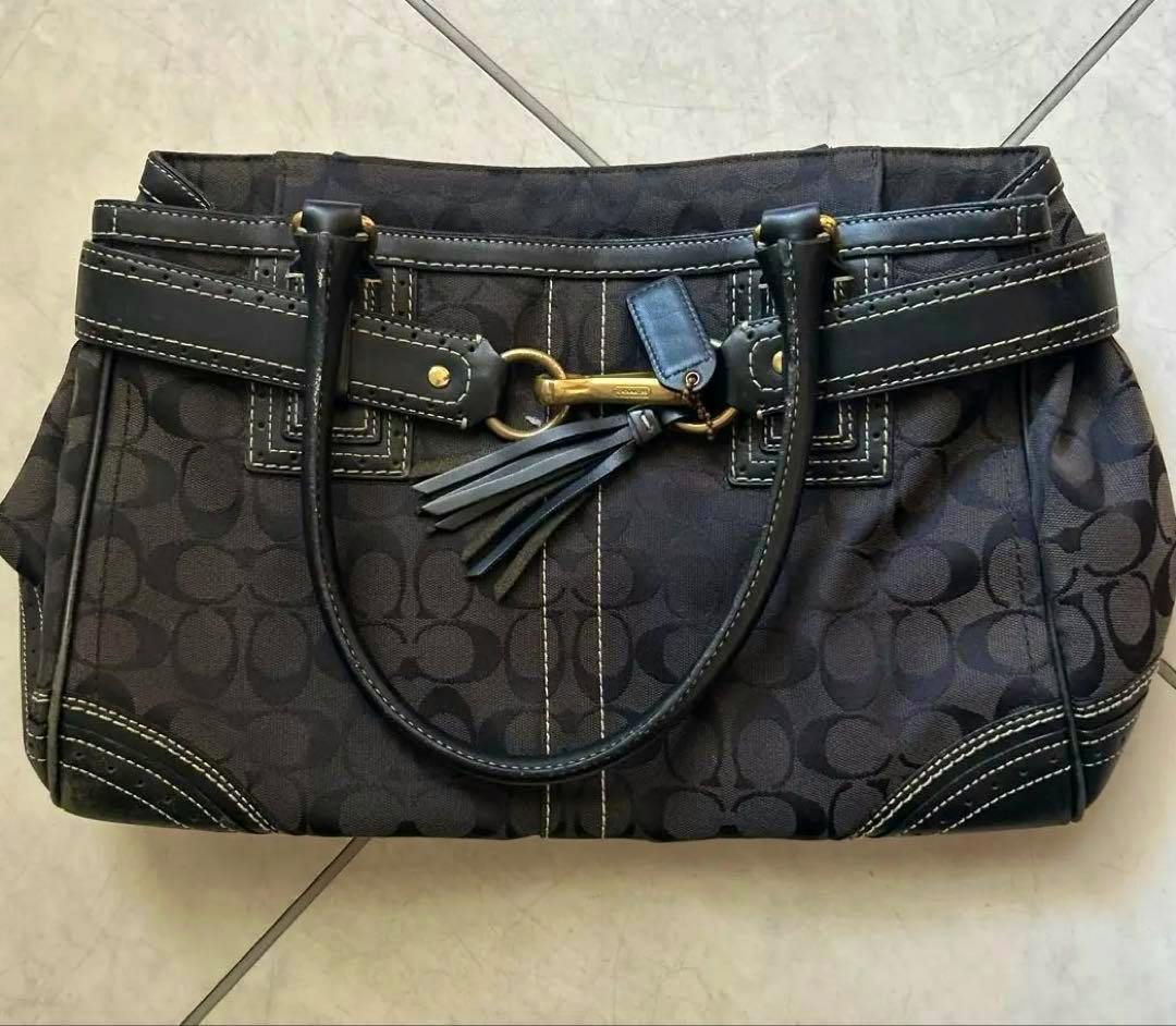 Black COACH signature-pattern handbag shown front-facing with leather handles, a tassel charm, and contrast stitching on a tiled floor.