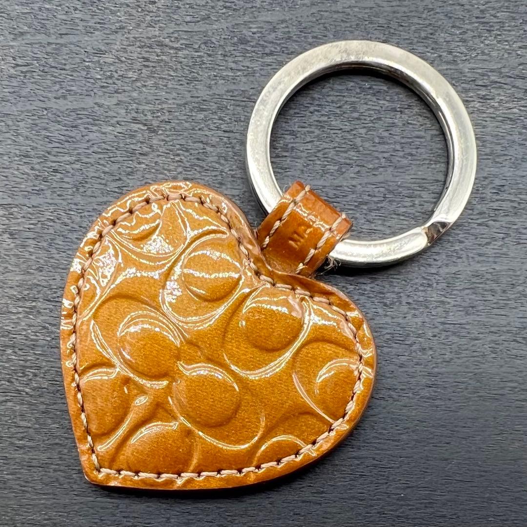 Heart-shaped COACH keyring with embossed signature pattern and a silver key ring on a dark background.