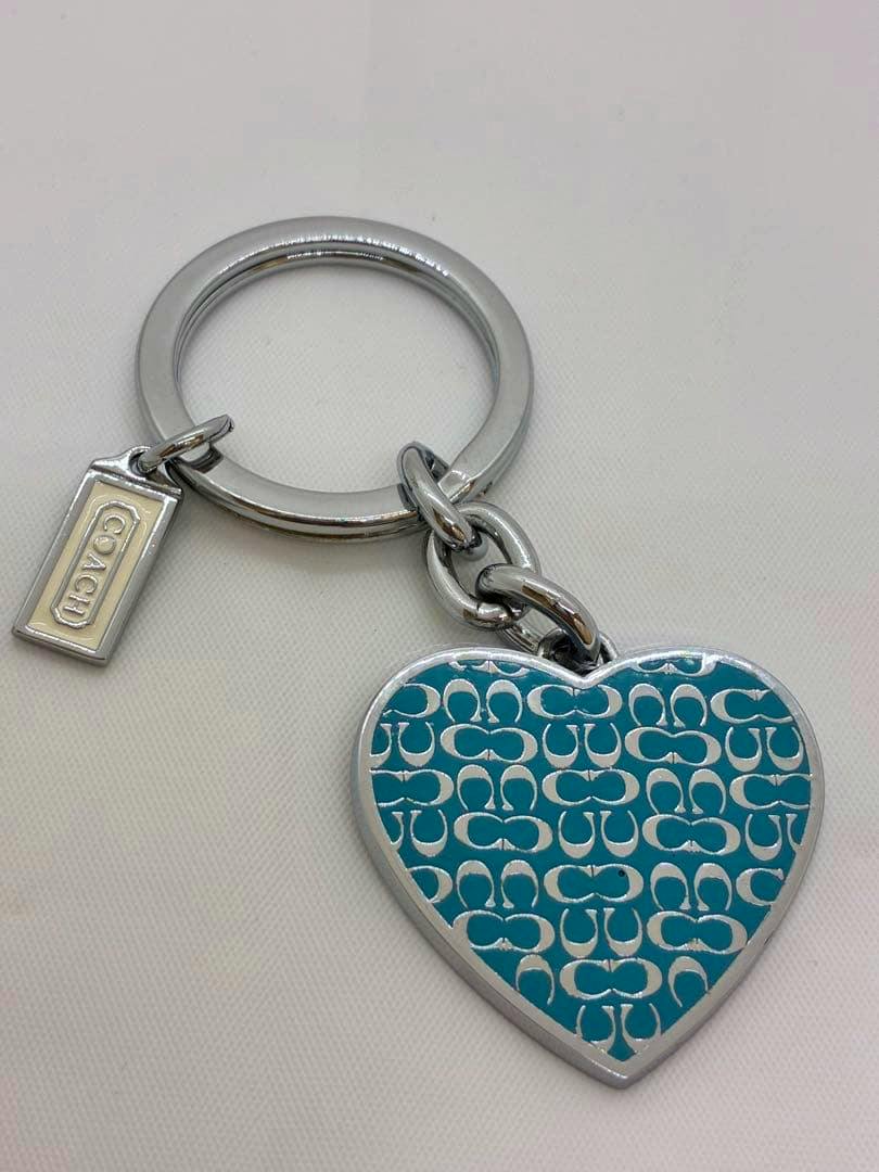 Blue heart-shaped COACH keychain charm with signature pattern attached to a metal key ring and small COACH tag.