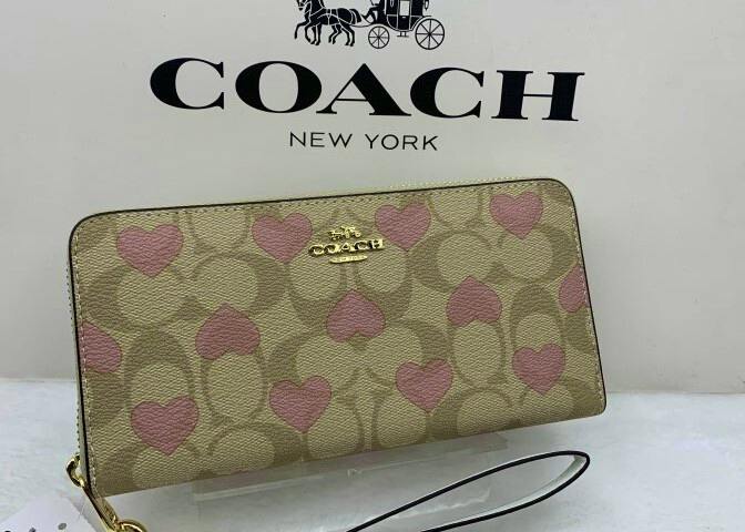 Front view of a COACH long zip wallet showing a beige signature pattern with pink heart print, gold COACH logo, and an attached wrist strap.