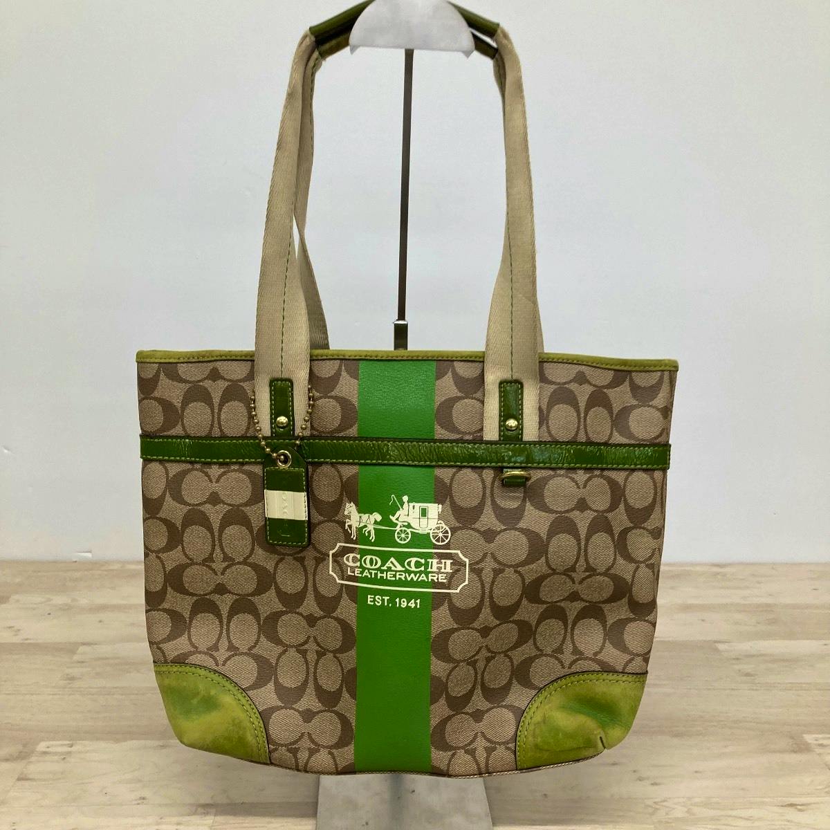 Front view of the COACH brown signature canvas tote bag with a central green stripe, horse-and-carriage logo, green trim, and beige handles.