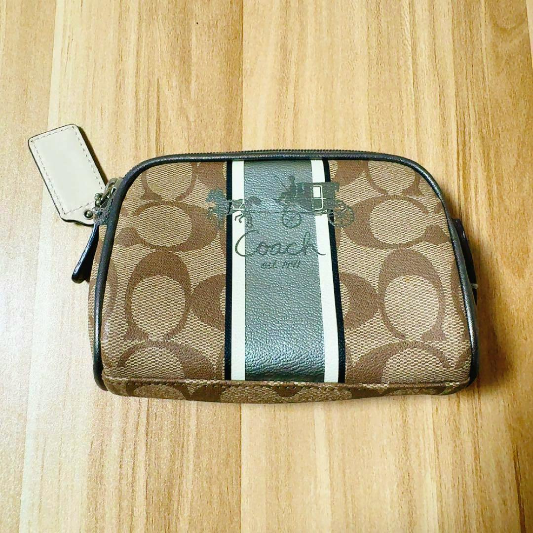 Brown Coach signature pouch with a central black-and-white vertical stripe and an attached tag, photographed on a wooden surface.