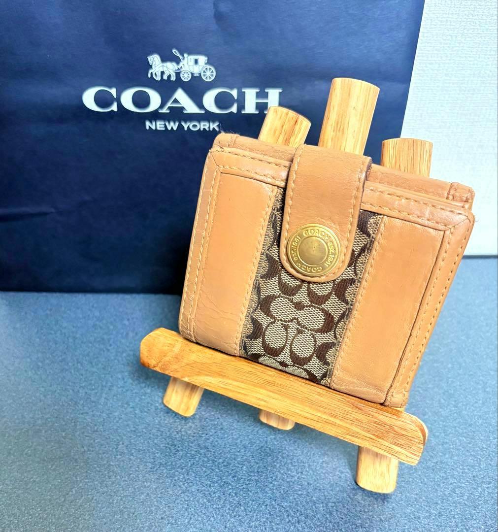 Light brown COACH signature leather bi-fold wallet shown on a small wooden stand with a COACH shopping bag behind it.