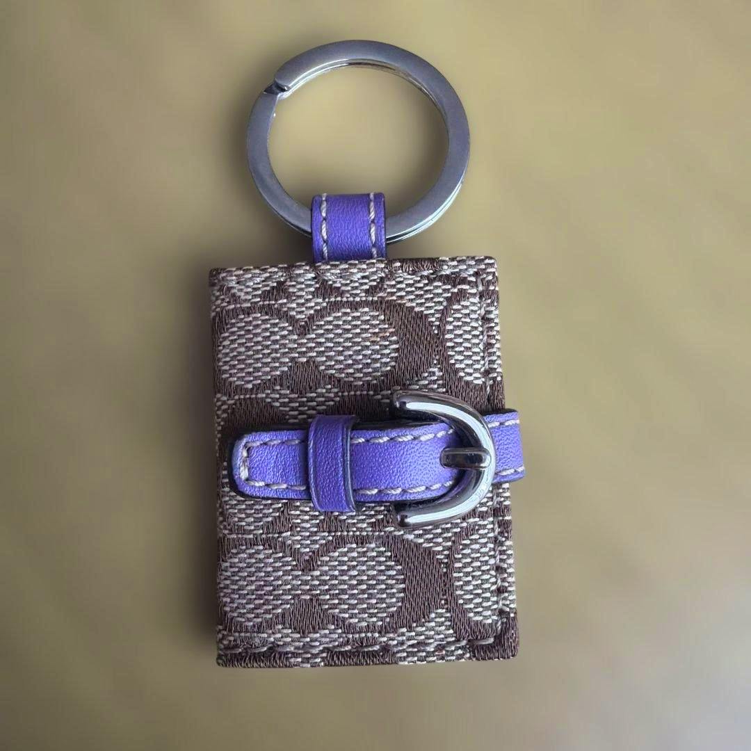 Brown Coach signature-pattern fabric keychain with a purple leather strap, silver buckle, and metal key ring.