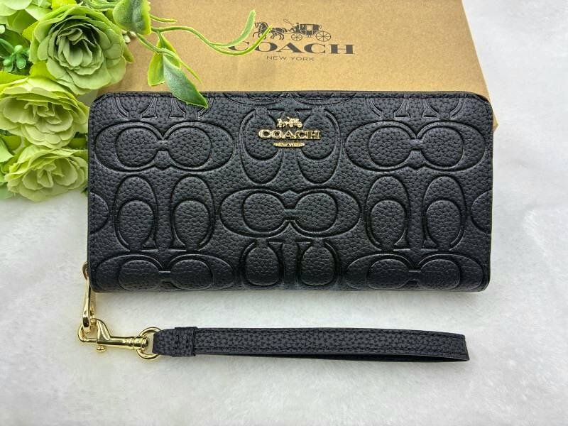 Black Coach long zip-around wallet with embossed signature pattern and a detachable wrist strap placed in front of a Coach box.