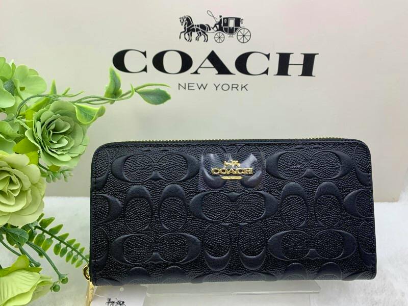 Black Coach zip-around long wallet with embossed signature pattern and gold logo plaque shown against a Coach backdrop.