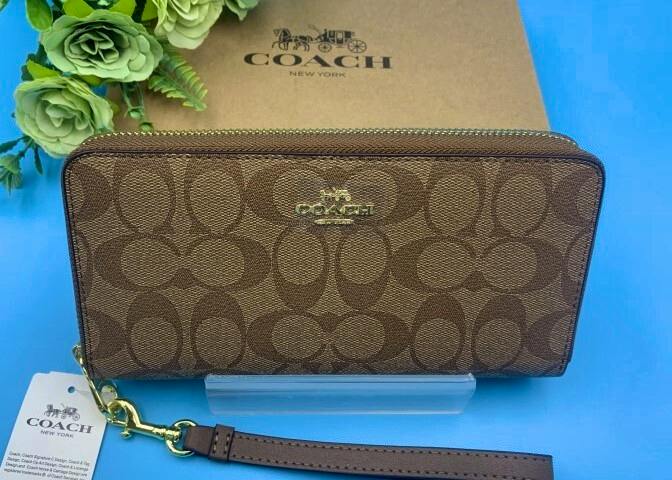 Brown Coach signature long zip-around wallet with gold logo and detachable wrist strap displayed with a Coach box and tag on a blue background.