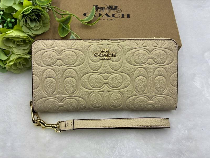 Beige COACH long wallet with embossed signature pattern and a detachable wrist strap shown on a white surface with a COACH box behind it.