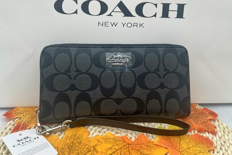 Front view of a black COACH signature long zip-around wallet with wrist strap in front of a Coach box.