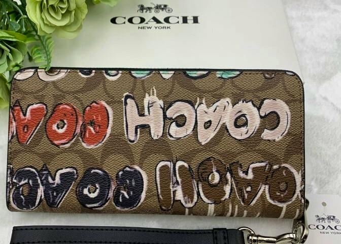A brown Coach signature canvas long zip-around wallet with graffiti-style "COACH" print laid flat with a Coach box and green flowers behind it.