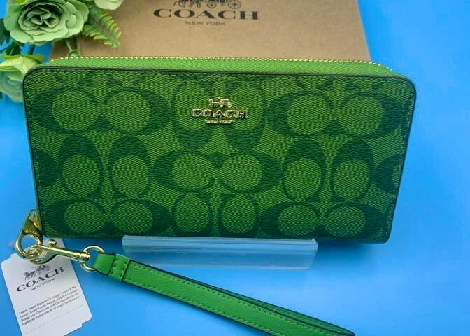 Green COACH long zip wallet with signature C pattern and gold logo, shown with a detachable wrist strap on a blue background.
