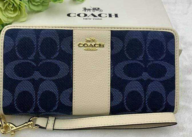 Front view of a navy Coach canvas wallet with a cream leather center strip, gold Coach logo, and attached wrist strap placed on a Coach box.