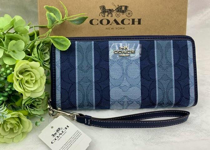 Blue Coach signature canvas long zip wallet shown front-facing with wrist strap, tag, and a Coach box behind it.