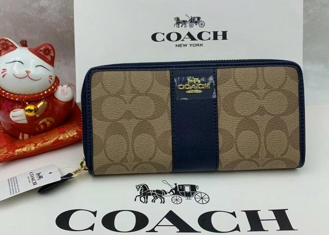 COACH long zip-around signature wallet in tan signature pattern with navy trim, displayed in front of a COACH box and a decorative lucky cat.