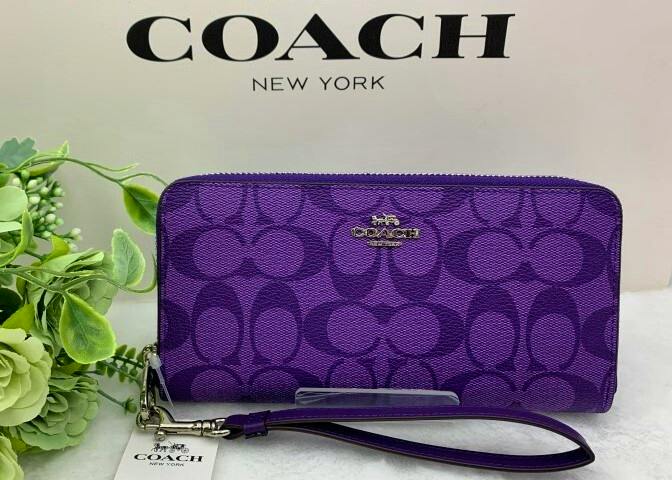Purple Coach long zip wallet with signature pattern and wrist strap displayed in front of a COACH New York backdrop.