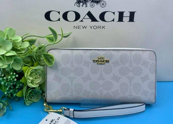 White COACH long zip canvas wallet with wrist strap displayed in front of a COACH box and decorative greenery.