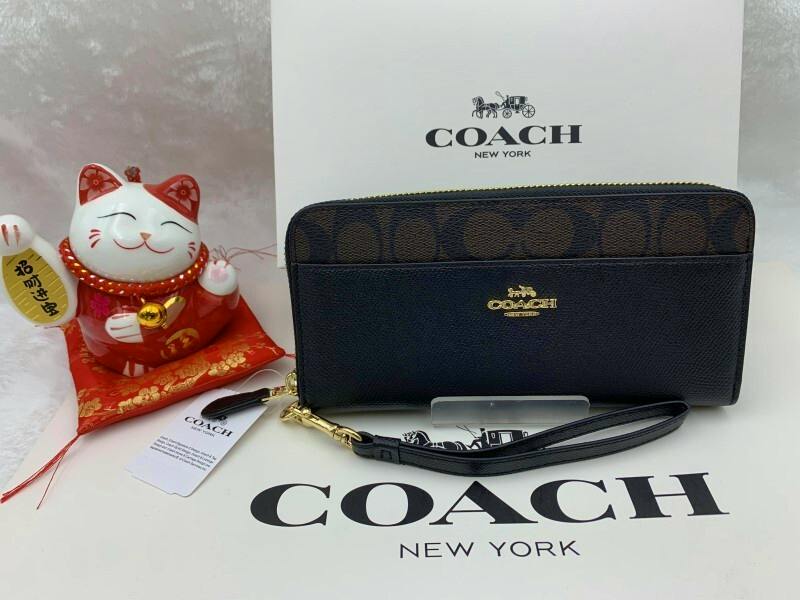 Front view of a COACH long zip men's wallet with signature print and wrist strap displayed on a COACH box.