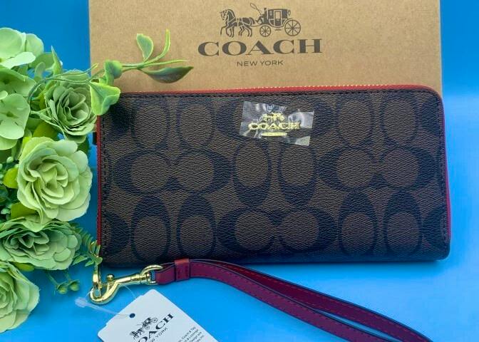 Coach signature long zip wallet with red wrist strap shown front-facing on a blue surface in front of a Coach box.