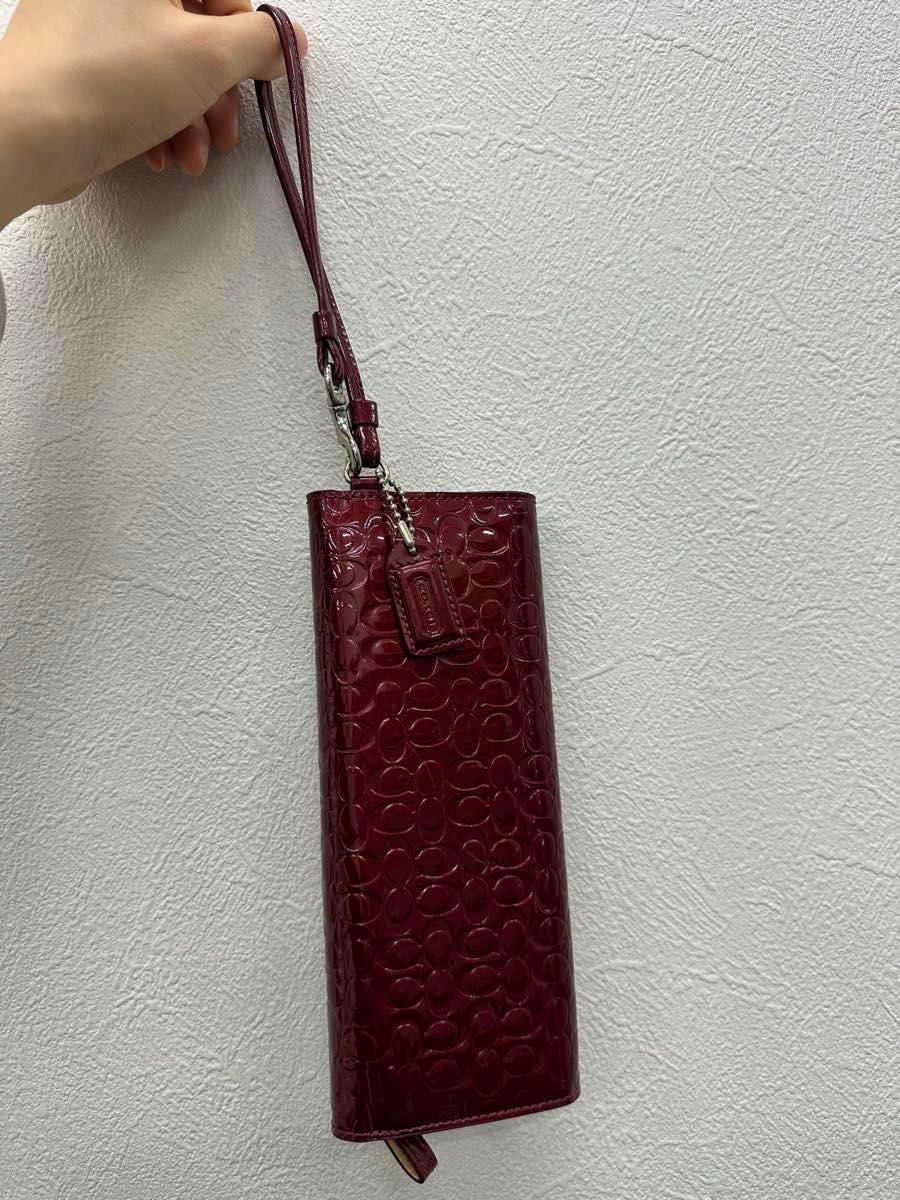 Red patent-leather long wallet with Coach signature pattern and wrist strap held against a white textured wall.