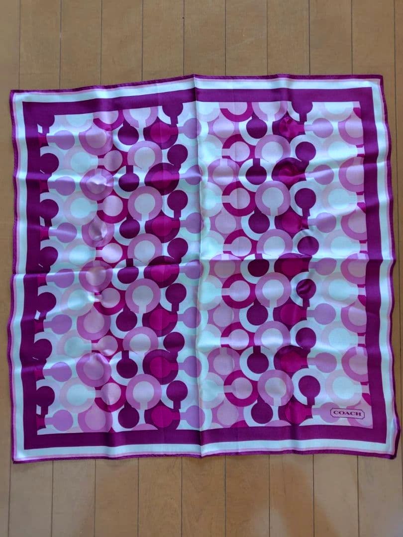 Pink COACH signature-pattern silk scarf laid flat on a wooden floor showing the repeating circular motif and border.