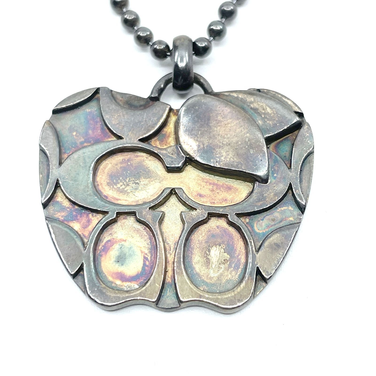 Close-up of the COACH signature pendant on a ball chain showing the metal surface and pattern.